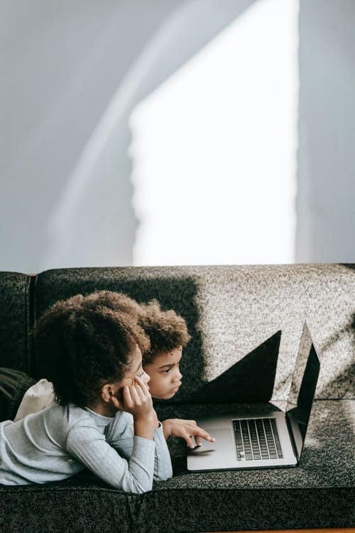 Two kids attending online classes while sitting on a couch in their home