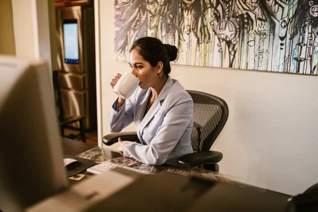 A woman drinks coffee from a cup while looking at her computer screen