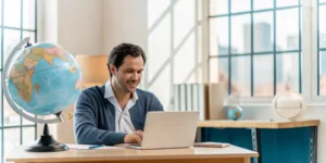 Man with a laptop and globe researches how to enroll in an accredited American online school abroad.